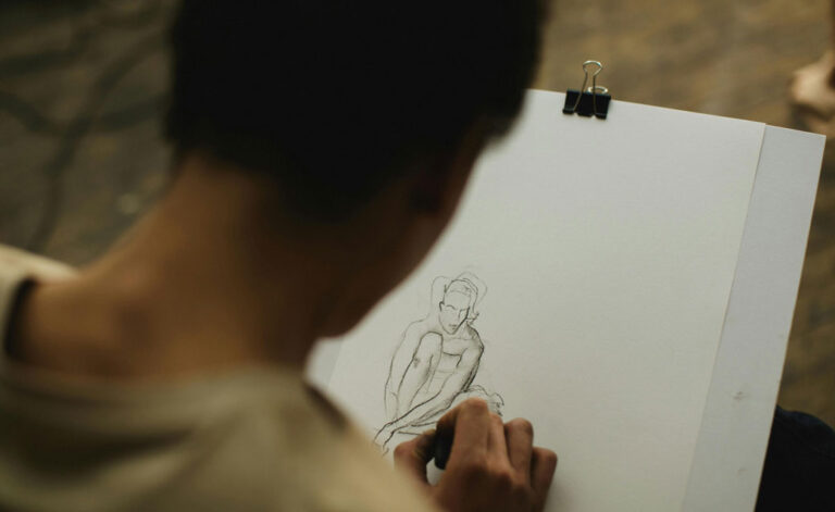 A stock photo image of a woman seen from behind, drawing a person on her sketchpad with a pencil