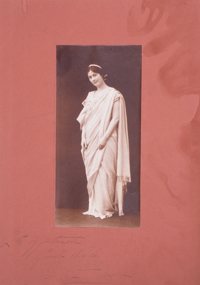 In the photograph "Isadora Duncan in Munich," the dancer softly smiles at the camera while wearing a light-colored draped dress.
