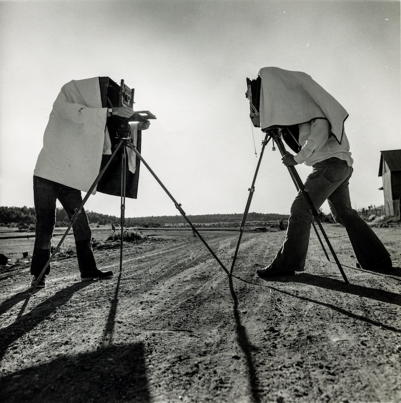 Arthur Tress' photograph "Camera Club Outing, Santa Fe, NM" depicts two people looking through their cameras while standing under dark cloths and facing opposite directions.