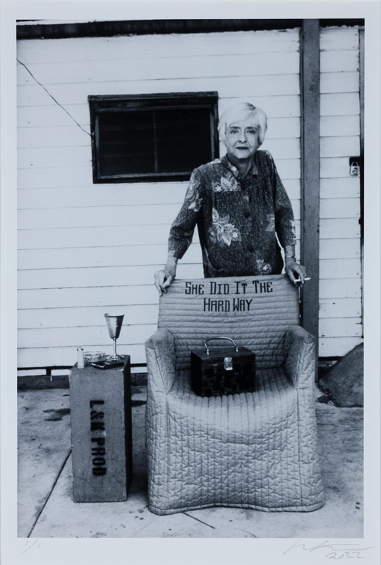 A black-and-white photograph of the actress Bette Davis by Nancy Ellison. The elderly Davis stands behind a chair with a slipcover that bears the words "She did it the hard way."