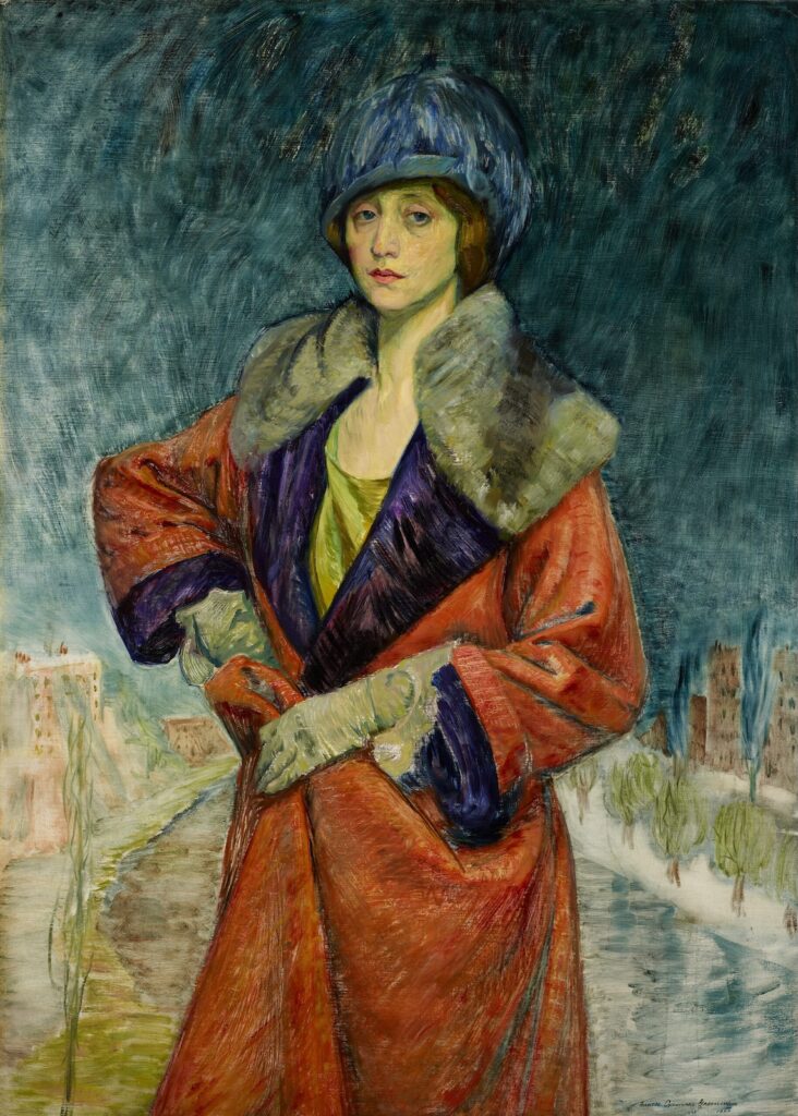 A self-portrait by artist Frances Cranmer Greenman from 1923. She shows herself almost full length, with both her hands on her right hip. She wears an orangey-red long coat with a fur collar, gloves and a blue hat.