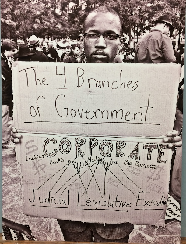 Charles Jerry Ayers' photograph on Plexiglass "Zucotti Park" depicts an Occupy Wall Street demonstrator who holds a sign that says "The 4 Branches of Government: Corporate Lobbies, Banks, Media, Big Business" as an illustration of hands holds puppet strings on "Judicial, Legislative, Executive"