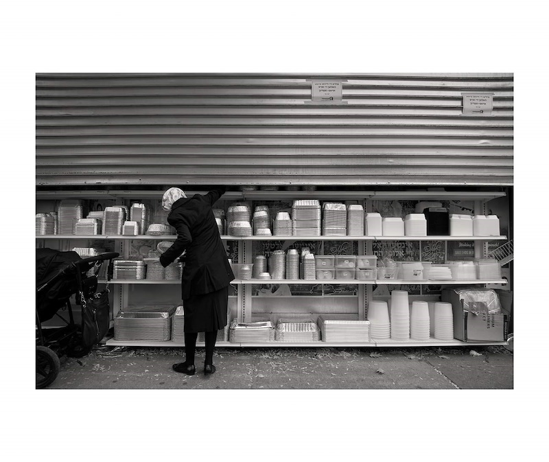 William Castellana's photograph "Woman Picking Food Container/ Penn St., Brooklyn, NY" shows the back of a woman in front of three shelves of aluminum and Styrofoam food containers.