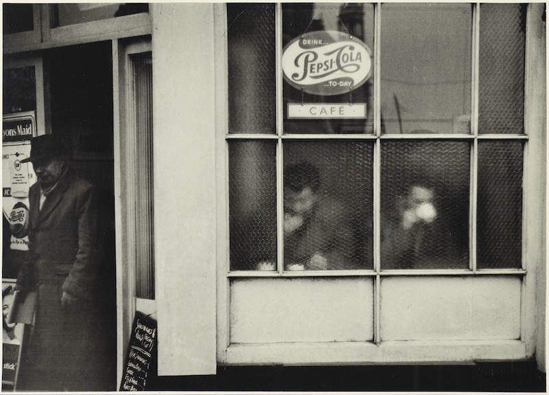 Lewis Morley's photograph "Café, London" depicts the exterior of a café. Two men eating and drinking can be seen through the window as another man exits through the door.