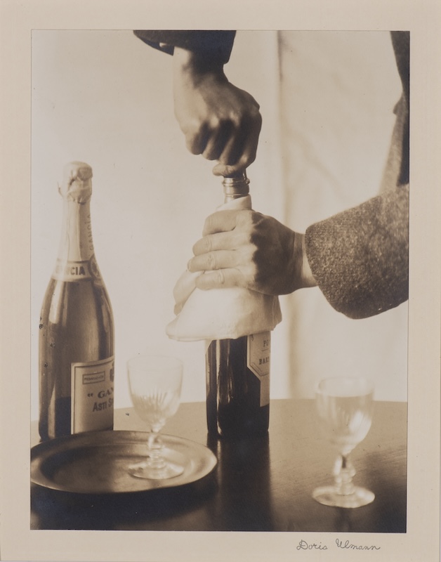 Doris Ulmann's photograph "John Jacob Niles Opening a Bottle of Wine" depicts two bottles of wine and two glasses on a table. A pair of hands comes from outside the frame to open one of the bottles.