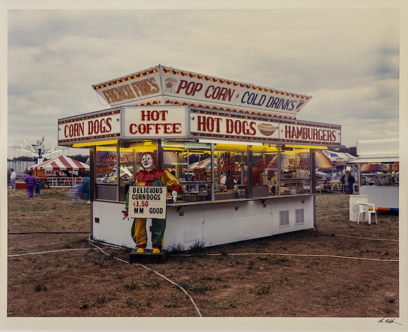 William Lee Wilson Jr.'s photograph depicts a mobile food stand at a county fair that sells hot dogs, hamburgers, pop corn, french fries and drinks. A smiling clown figure holds a sign that says "DELICIOUS CORNDOGS $1.50 MM GOOD."