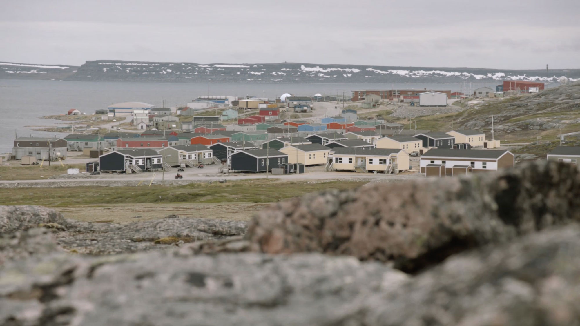 A screenshot from asinnajaq's film "Three Thousand" that shows low houses clustered together near the water, seen from a distance.