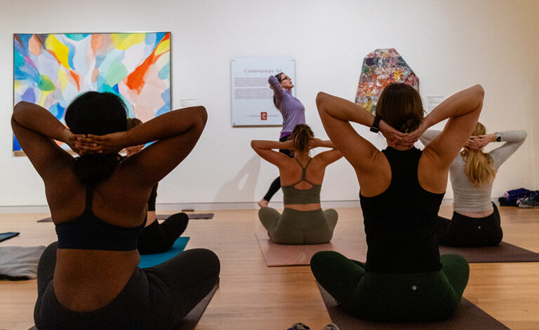 Five women are seated in the gallery practicing yoga. Two artworks can be seen on both sides of the instructor.