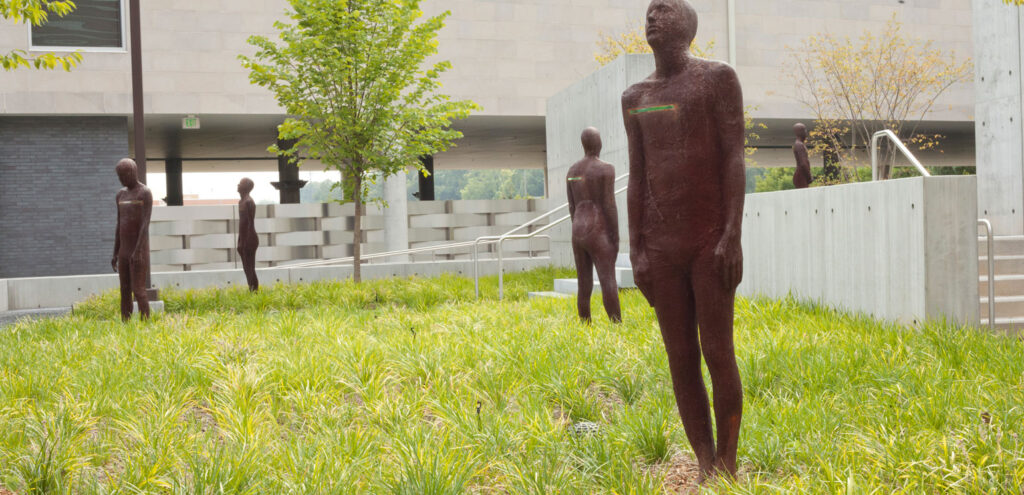 An installation photograph showing Steinnun Thorarinsdottir's sculptures in the exhibition "Horizons" at the Georgia Museum of Art. Made of rusted steel, they look like and are about the size of human figures, standing with their arms by their sides and each embedded with a strip of glass at about chest height.