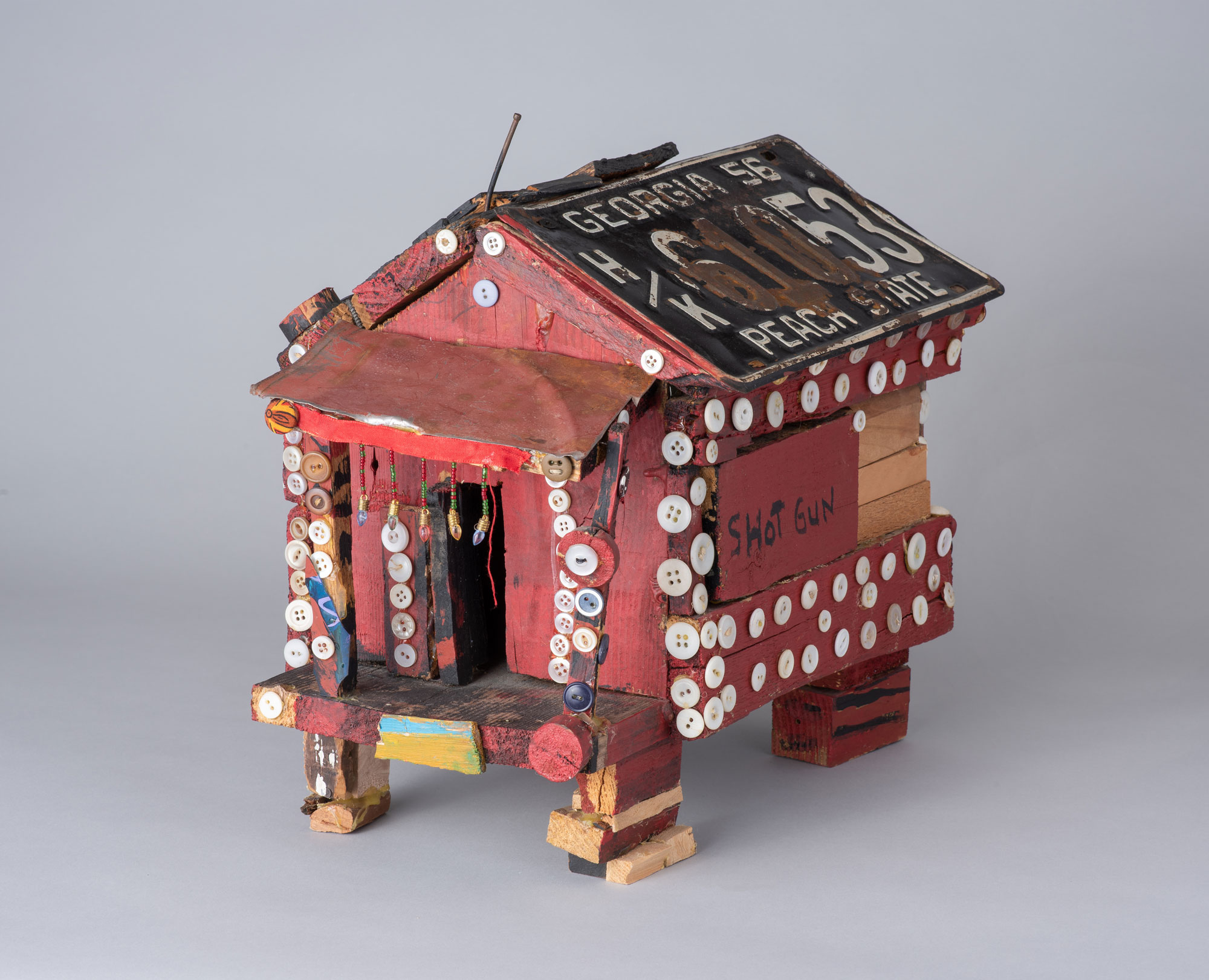 A small sculpture of a shotgun shack by Beverly Buchanen, painted a winey red and adorned with buttons. Two license plates form the roof.