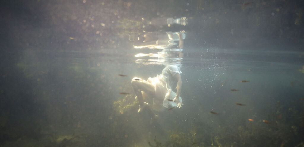 a detail of a photograph by Allison Janae Hamilton that shows the author, from below, wearing a white dress and submerged in water