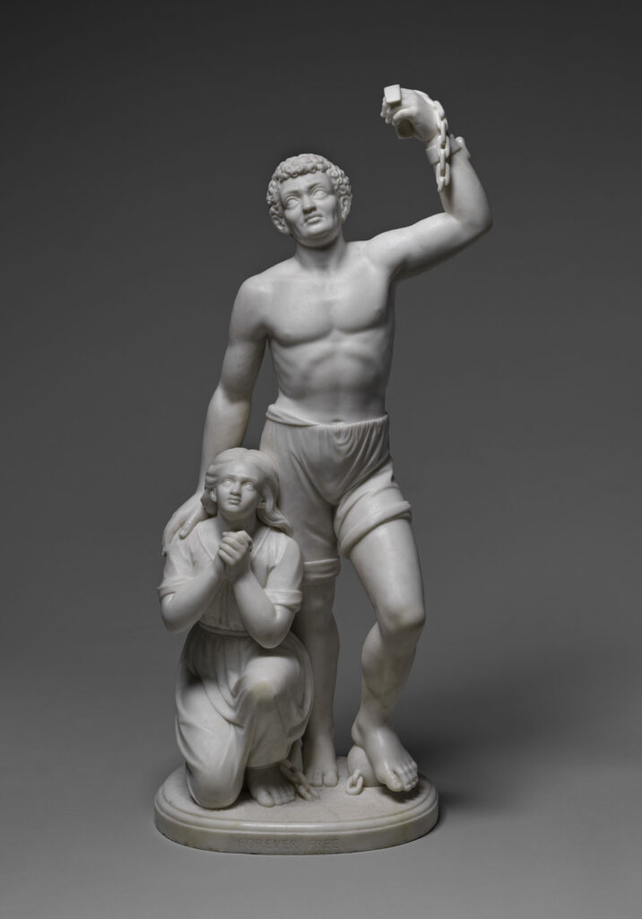 "Forever Free," a marble sculpture by Edmonia Lewis that shows a standing man, freed from enslavement, raising his left hand, around the wrist of which we can see a broken shackle. Kneeling at his side is a woman with her hands clasped in prayer.