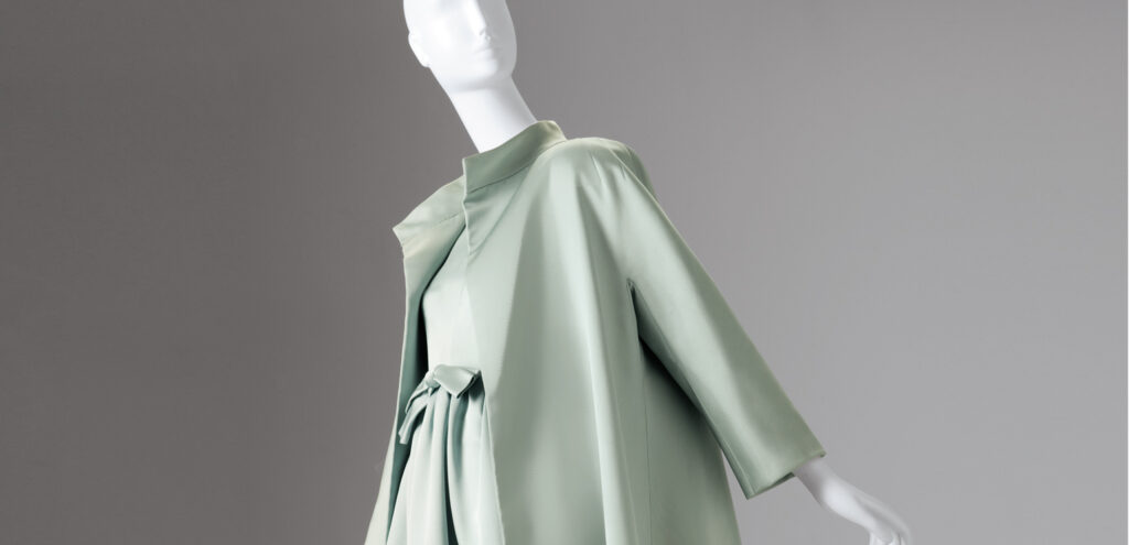 A color photograph of a mannequin wearing a pale green Balenciaga dress and a matching loose coat