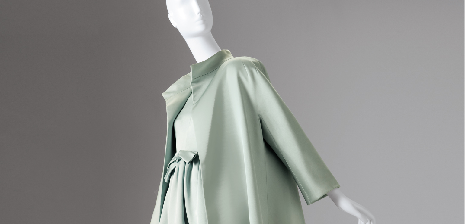 A color photograph of a mannequin wearing a pale green Balenciaga dress and a matching loose coat