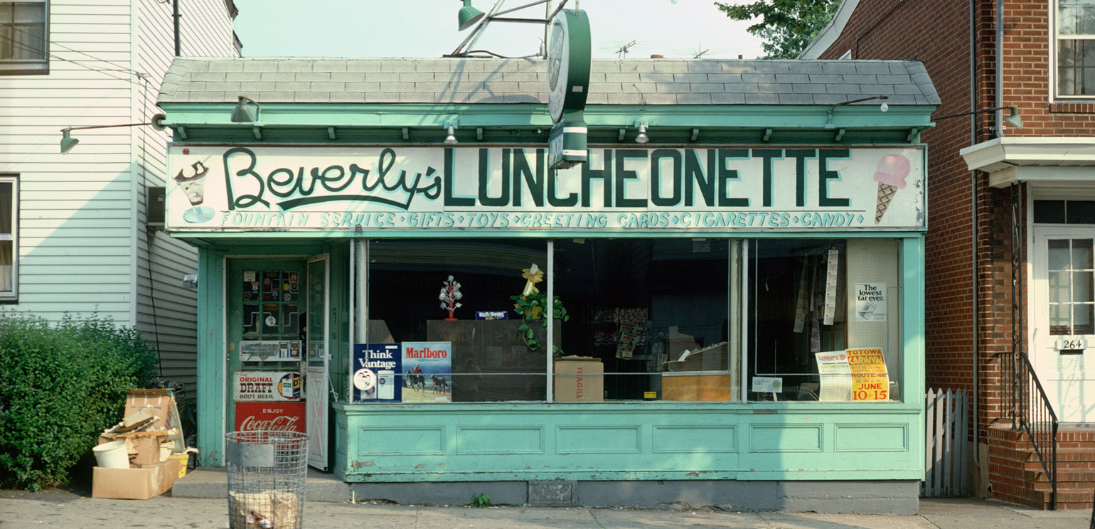 A horizontal color photograph by John Baeder of a pale greenish-blue-painted restaurant called Beverly's Luncheonette, seen from the outside.