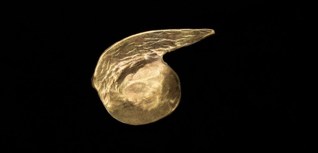 A piece of hammered gold jewelry by David Hayes that looks like a comma rotated 90 degrees counterclockwise, with the tail pointing to the right