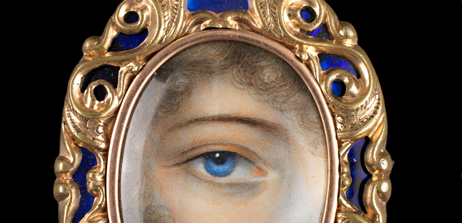 Close-up detail of a piece of eye jewelry, a gold-framed pendant with details in blue that features a close-up painting of a woman's blue eye