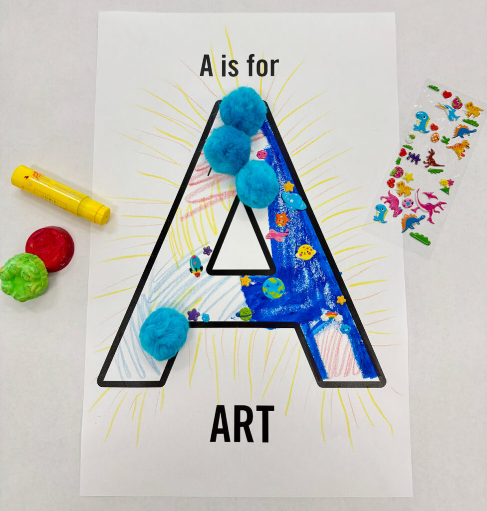 A printed outline of a capital letter A with the words "A is for ART." Someone has decorated the A with pompoms, markers, stickers and oil pastels.