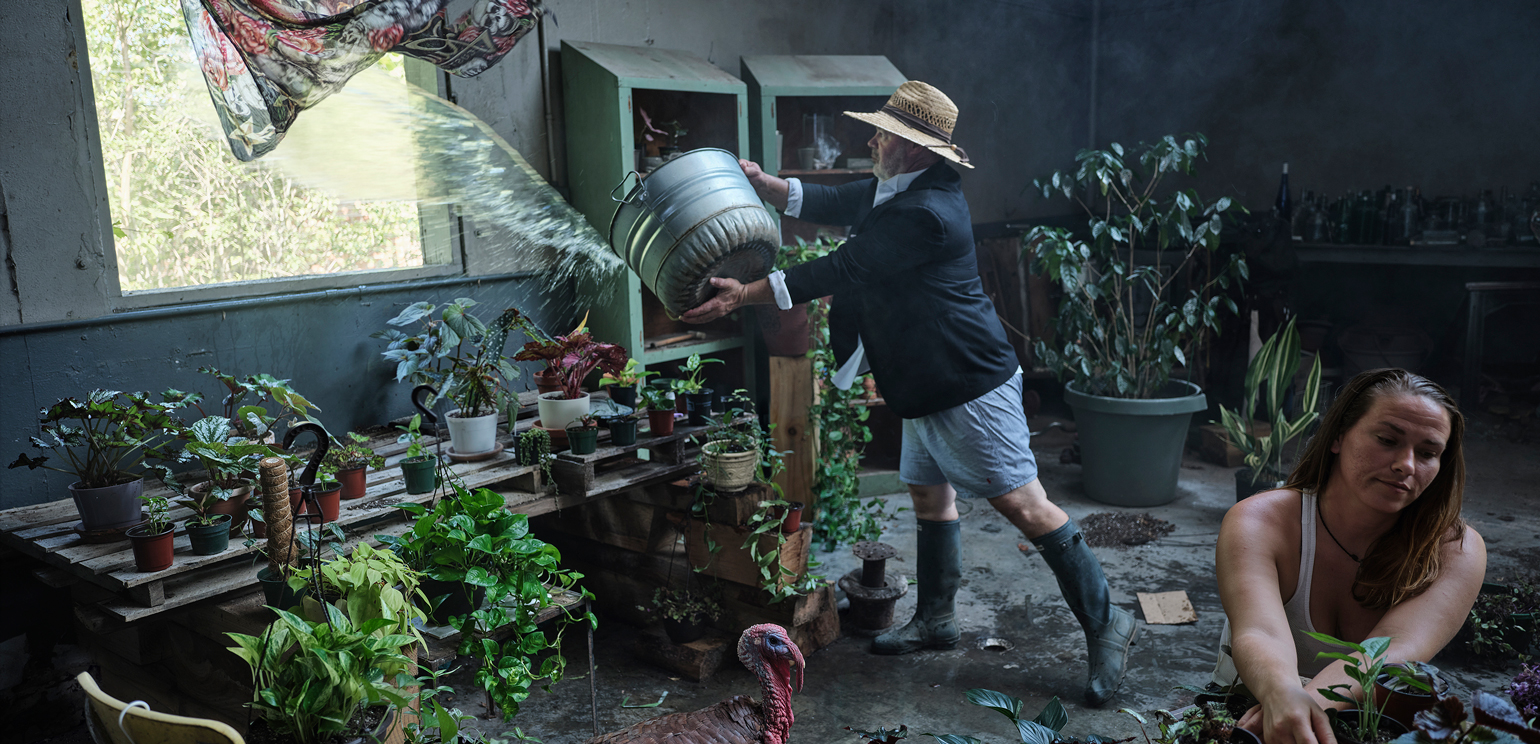 A detail of Jim Fiscus' photo "Judging Room." A bearded man in a hat and shorts throws a bucket of water out a window. A woman smokes as she repots plants. A turkey roams the room full of plants.
