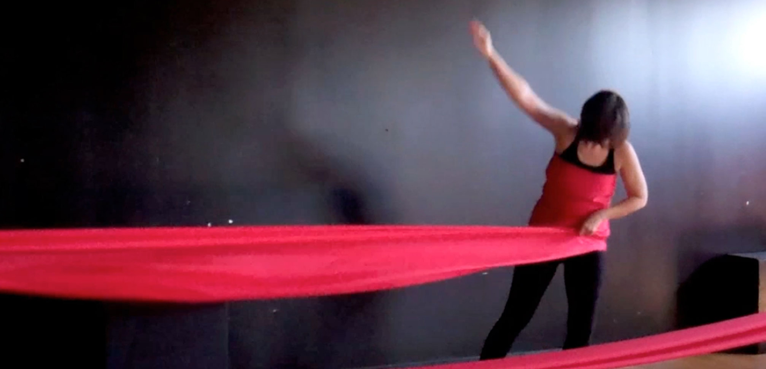A still from a video of a woman in a black unitard performing a dance with a long red piece of fabric. It's wrapped around her body twice, then continues off the screen before looping back.