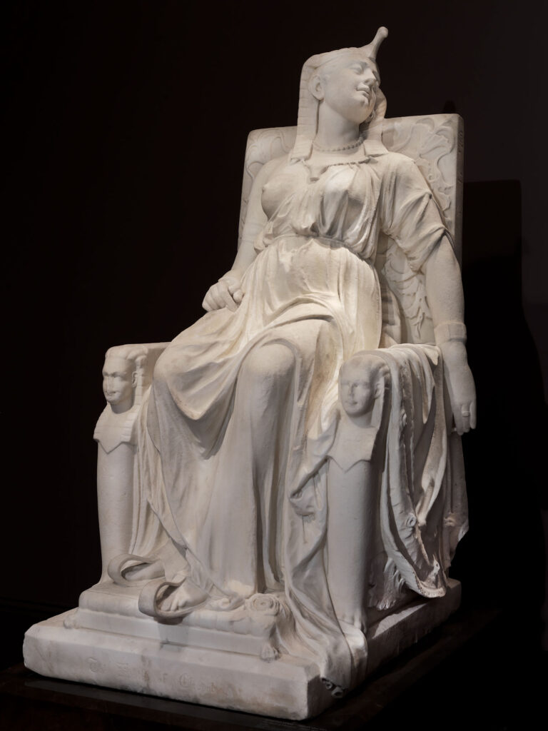 Edmonia Lewis portrayed Cleopatra in the moment after her death, wearing her royal attire, in majestic repose on a throne. The identical sphinx heads flanking the throne represent the twins she bore with Roman general Marc Antony, while the hieroglyphics on the side have no meaning.
