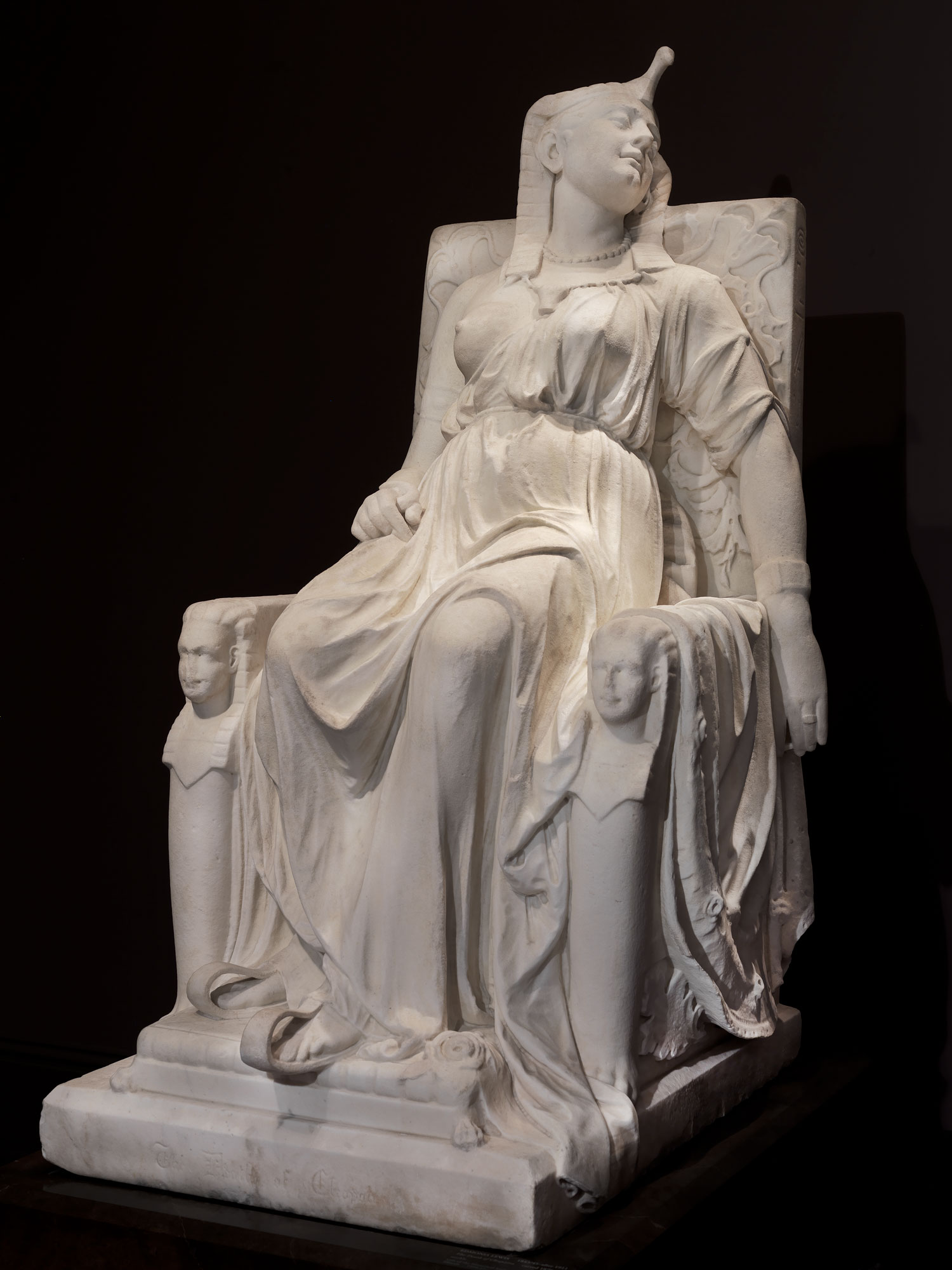 Edmonia Lewis portrayed Cleopatra in the moment after her death, wearing her royal attire, in majestic repose on a throne. The identical sphinx heads flanking the throne represent the twins she bore with Roman general Marc Antony, while the hieroglyphics on the side have no meaning.