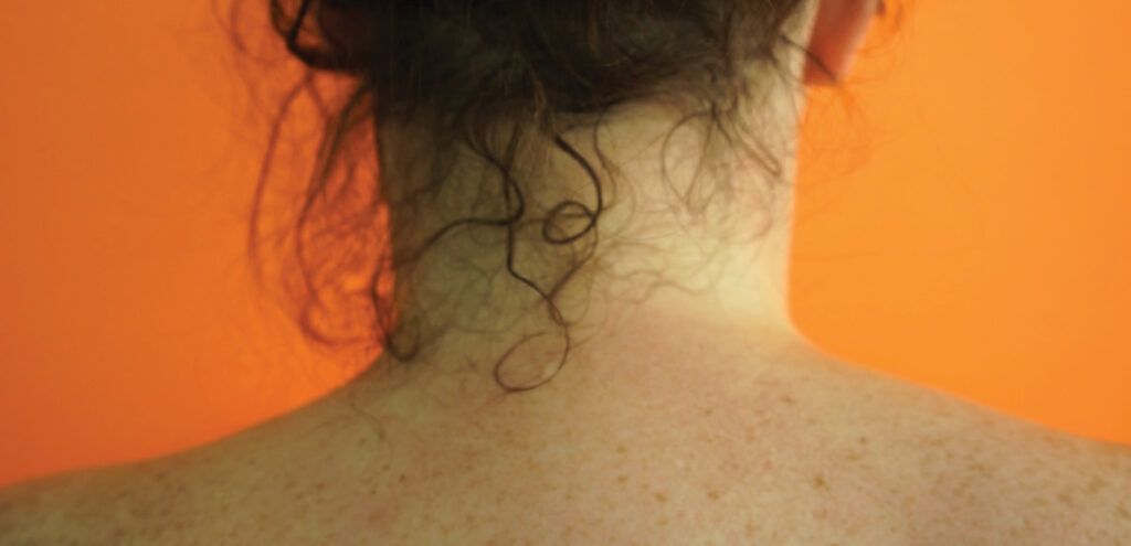 A detail of a still from a video by UGA MFA candidate Stephanie Sutton that shows the back of a woman's neck. Her shoulders are bare, her skin has freckles, a few tendrils of her curly dark hair escape its updo to touch her neck and the background is orange.