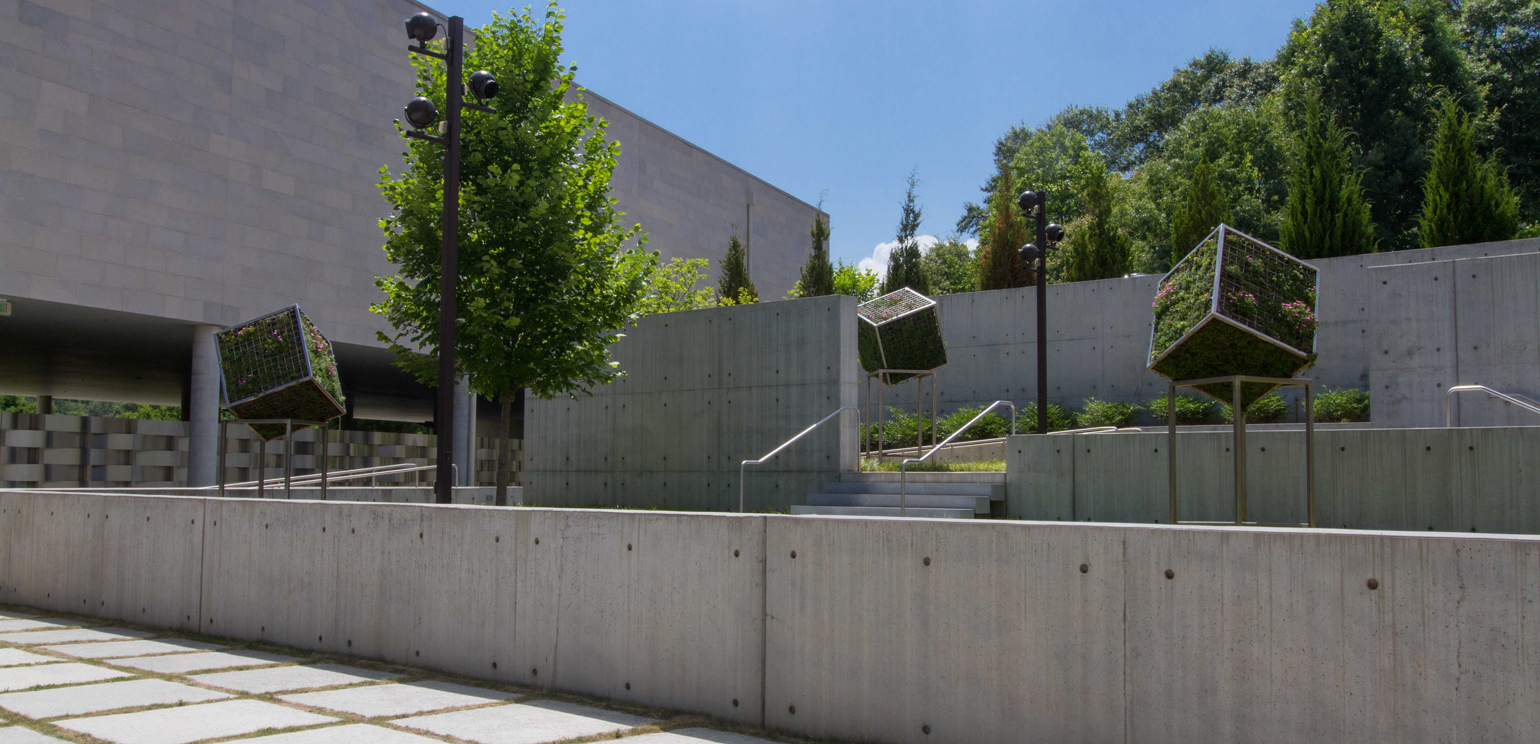 An installation photograph of the exhibition "Terra Verte" in the museum's sculpture garden. Cube-shaped steel cages mounted at an angle on metal legs hold live plants that grow through the mesh.