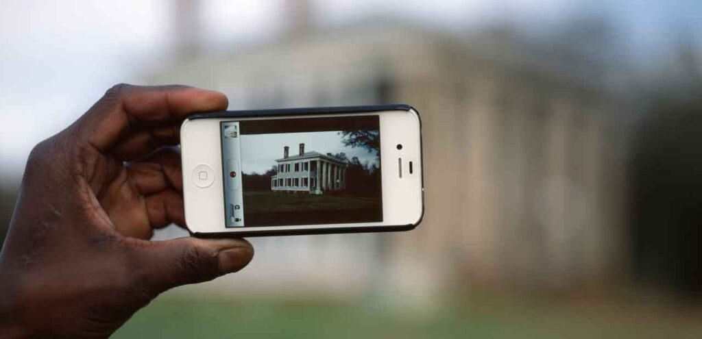 Detail of a photograph by Ramell Ross called "iHome." It shows a Black person's hand holding a phone with a photograph of a plantation on it, with the actual scene blurred out in the background.