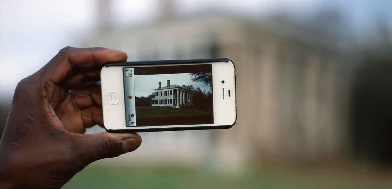 Detail of a photograph by Ramell Ross called "iHome." It shows a Black person's hand holding a phone with a photograph of a plantation on it, with the actual scene blurred out in the background.
