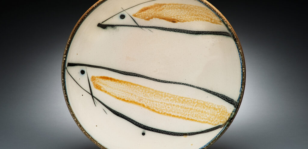 A ceramic plate made by the artist Michael Simon, in a color photograph looking down at the plate. It features two abstracted fish, rendered in black ink with a swath of yellow lengthwise down their bodies.