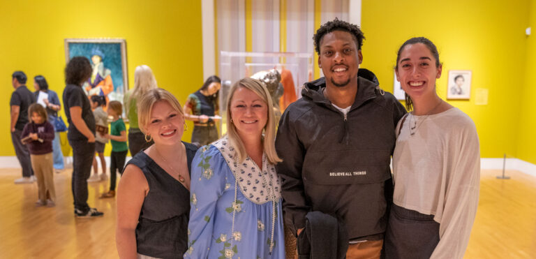 Four people standing side by side for a photo in the galleries. Behind them, other people mill about in the exhibition.