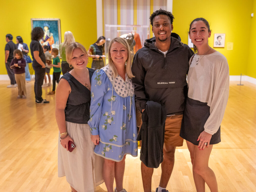 Four people standing side by side for a photo in the galleries. Behind them, other people mill about in the exhibition.
