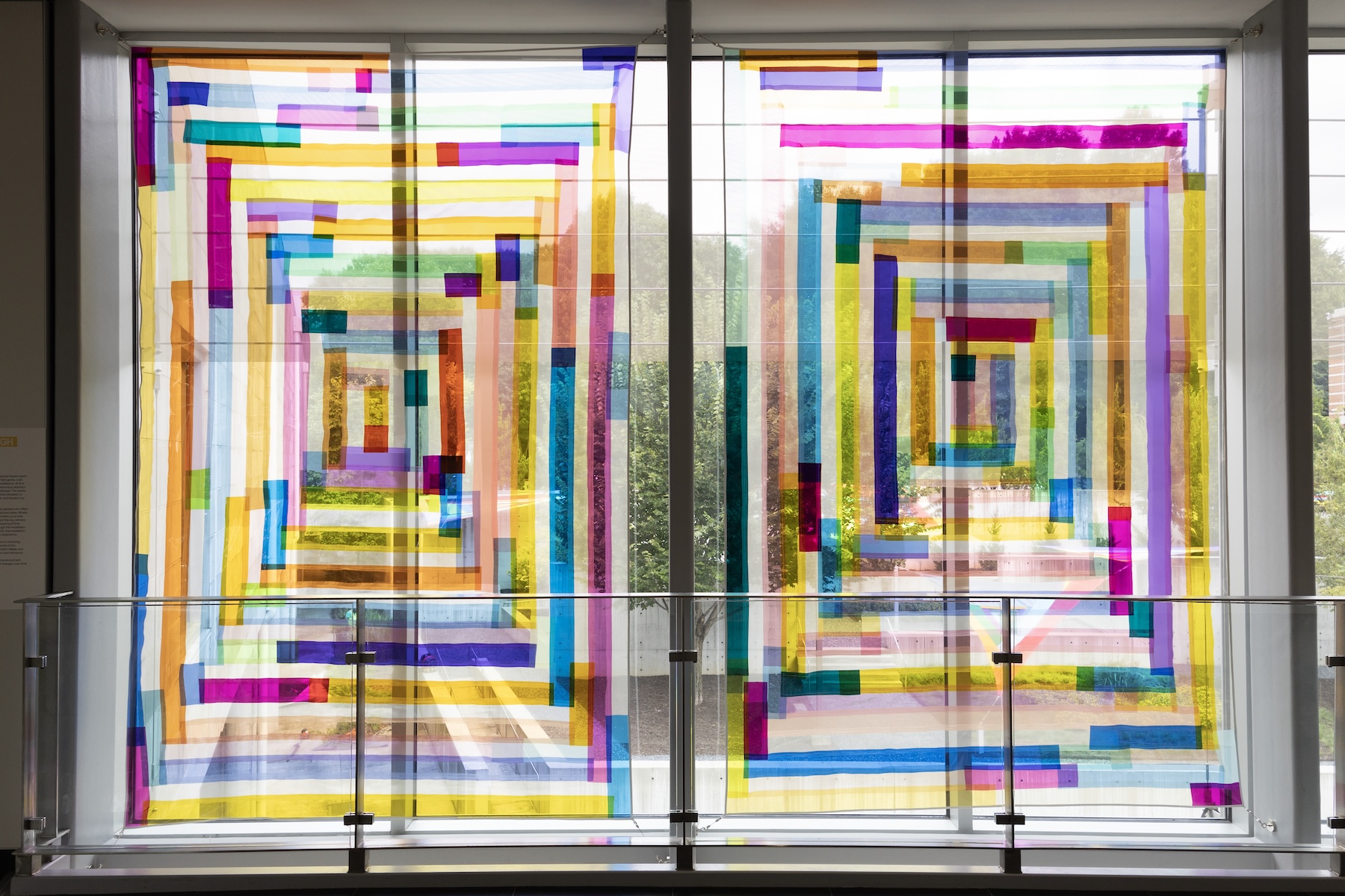 Two textile works made of translucent multi-colored strips hanging in the window. The sculpture garden can be seen in the background.