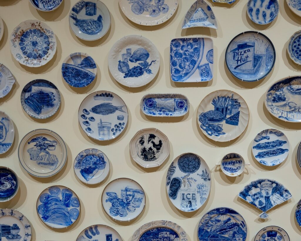 A collection of small plates hanging on the wall show blue paintings of last meals requested by incarcerated people on death row