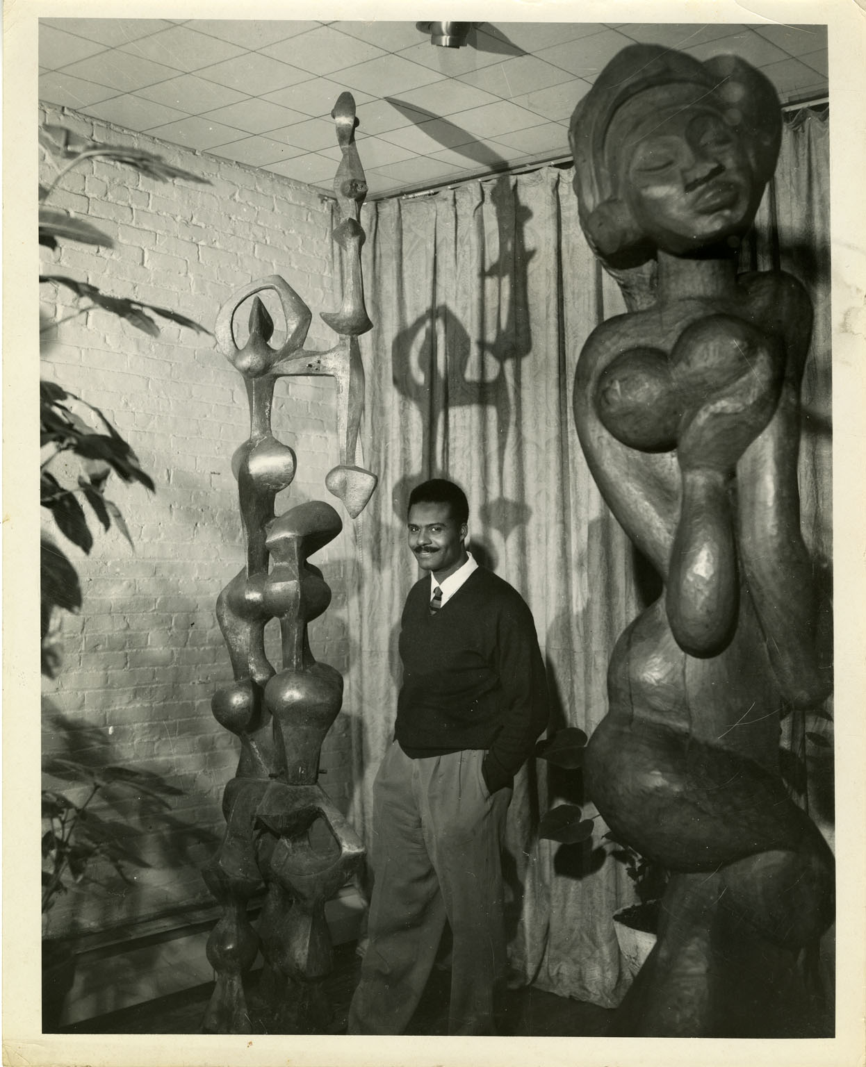 Black and white photograph of John Rhoden with his sculptures, including "The Offering," in the Rhoden home at 23 Cranberry Street in Brooklyn, New York.