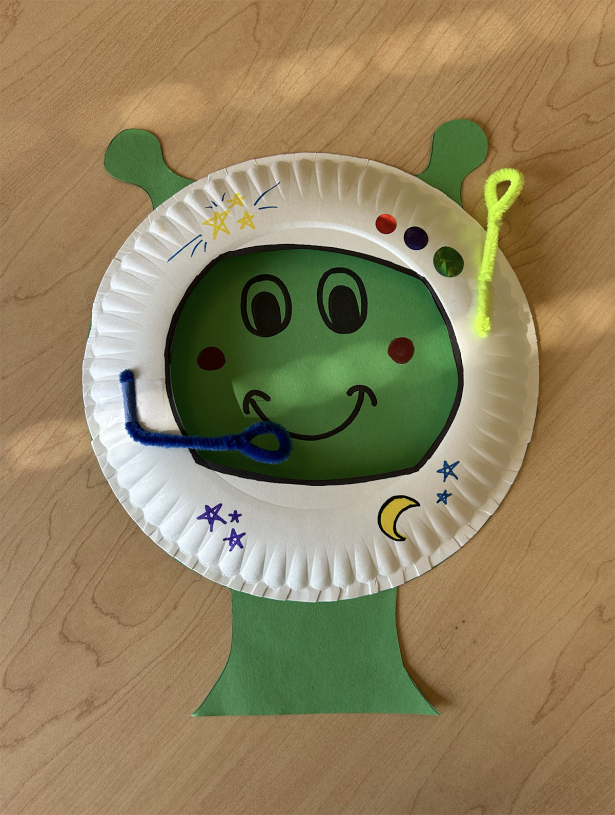 A green alien face made out of paper, wearing a helmet mask made out of a paper plate decorated with markers and pipe cleaners