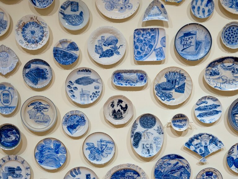 A collection of small plates hanging on the wall show blue paintings of last meals requested by incarcerated people on death row