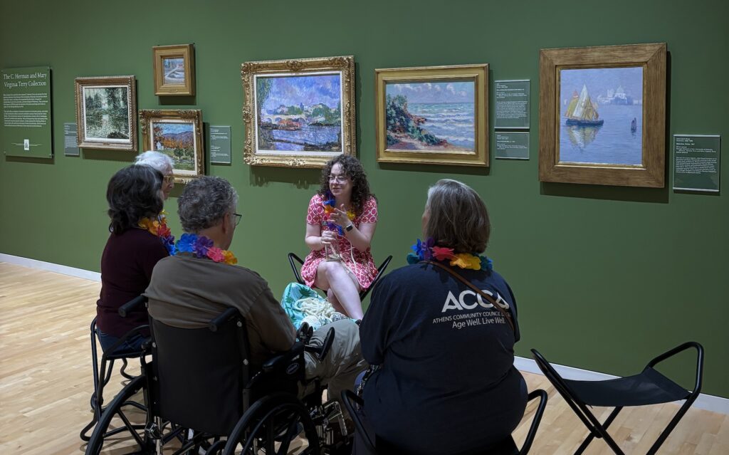A small group of visitors from the Athens Community Council on Aging listen to Mallory Lind, associate curator of community engagement and outreach, as she speaks about art. Multiple impressionist paintings hang on the wall behind her.