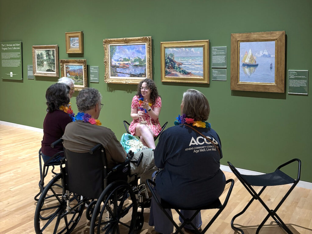 A small group of visitors from the Athens Community Council on Aging listen to Mallory Lind, associate curator of community engagement and outreach, as she speaks about art. Multiple impressionist paintings hang on the wall behind her.