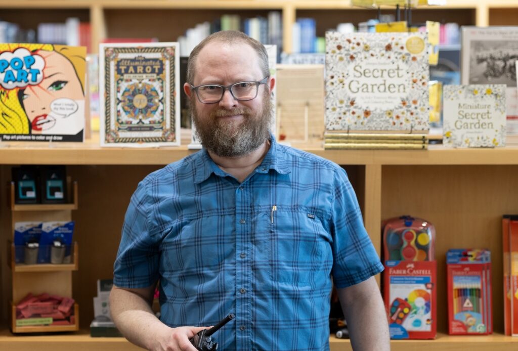 Adam Clark, manager of visitor experience, stands against a shelf displaying coloring books and art activities in the Museum Shop