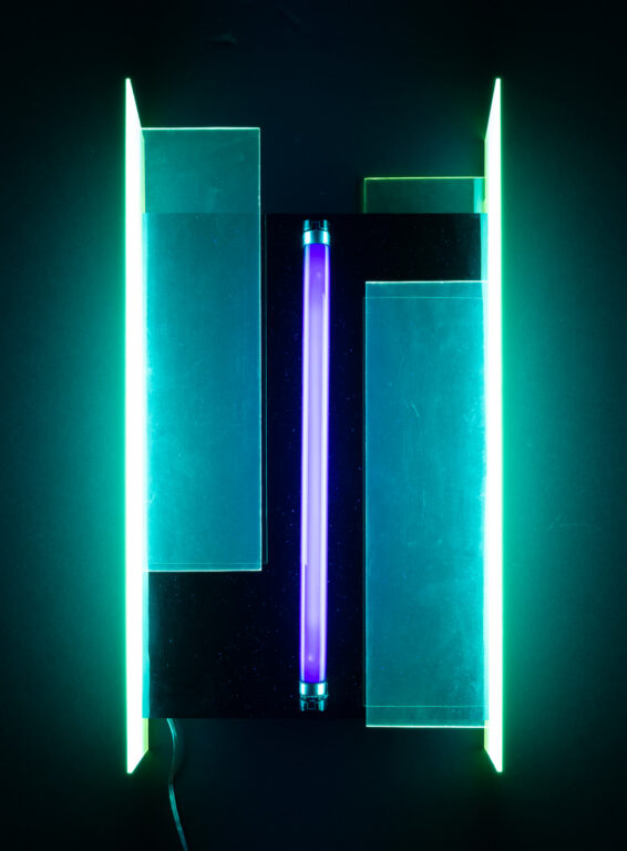 A light sculpture by Lila Katzen that features an ultraviolet light tube installed vertically between two sheets of acrylic. When plugged in and turned on, the acrylic appears to light up too, in two bright vertical bands of light green.