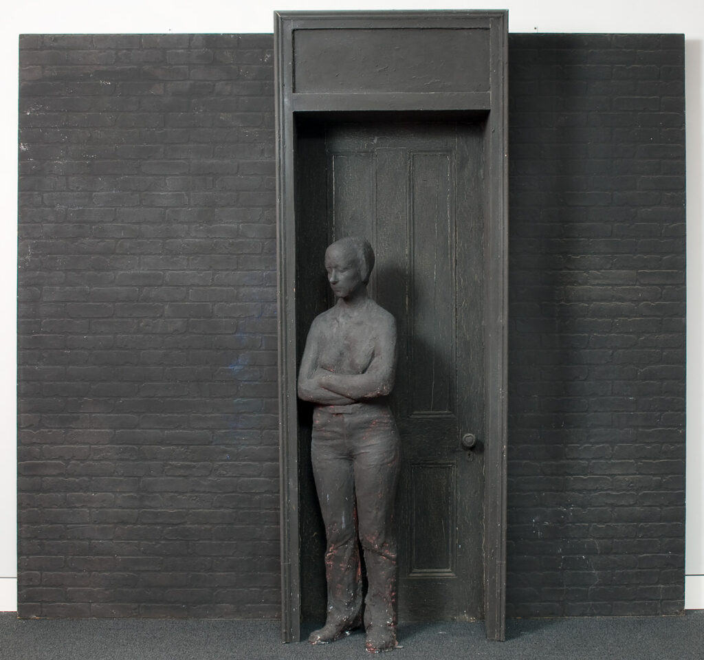 George Segal’s “Young Woman in Doorway” is a large sculpture that features a life-size woman leaning against a doorway. The door behind her is closed. Behind the doorway, which protrudes, is a wall of brick. The whole sculpture is made of plaster over wire and other bases and painted black.