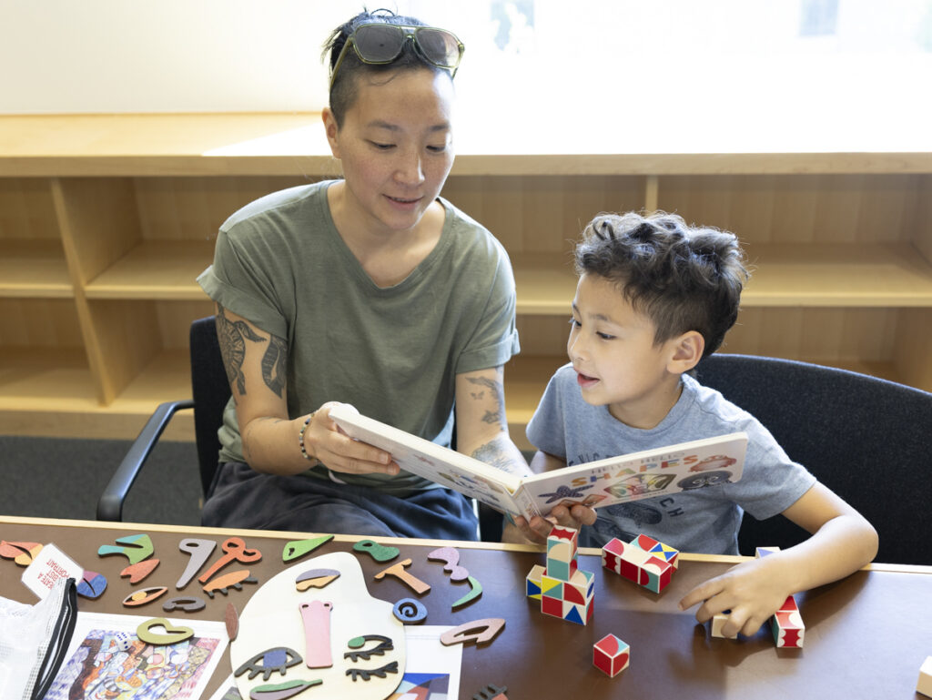 A mother shows her son a picture book about shapes. Various art activities are laid out on the table.
