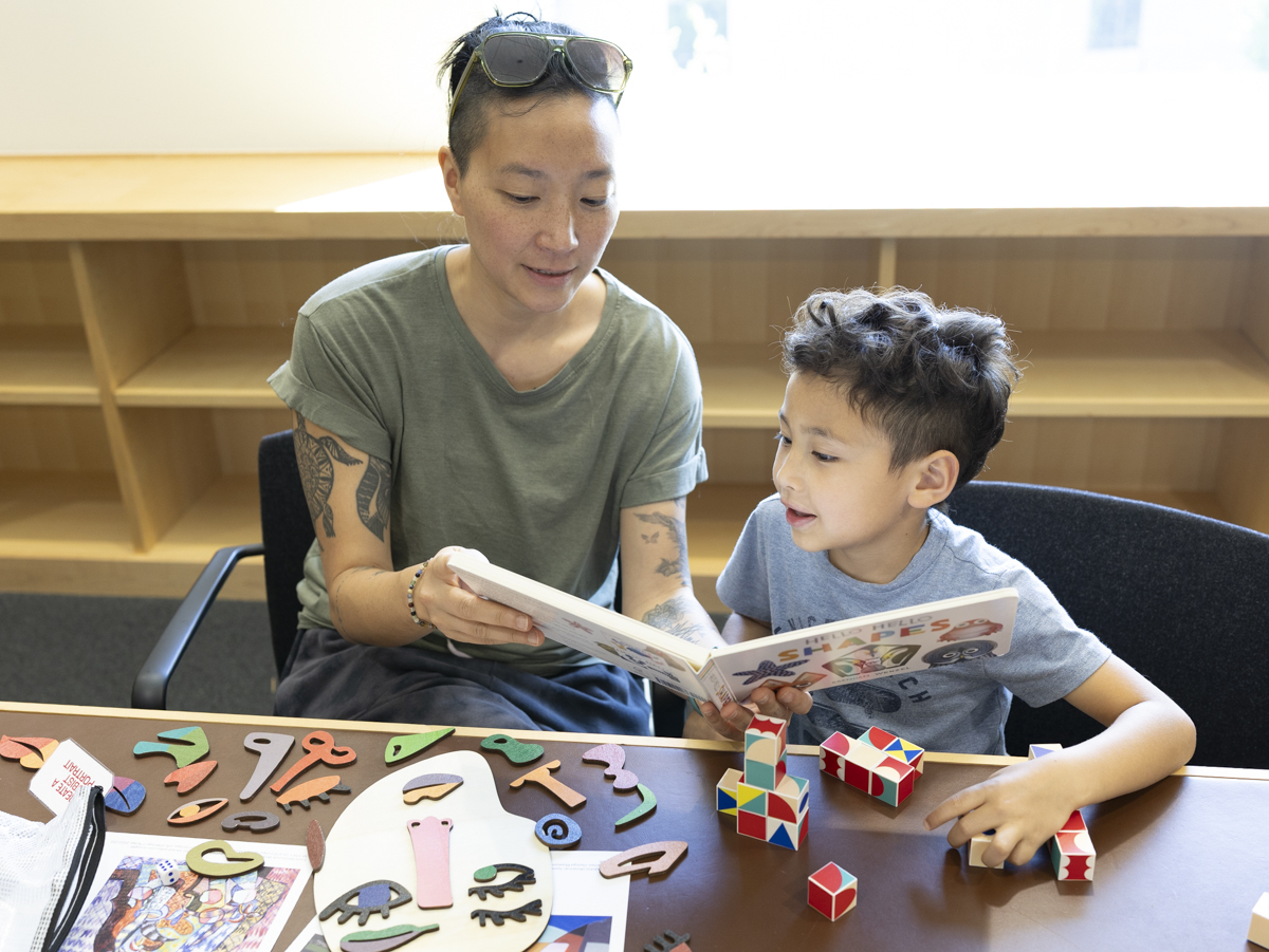 A mother shows her son a picture book about shapes. Various art activities are laid out on the table.