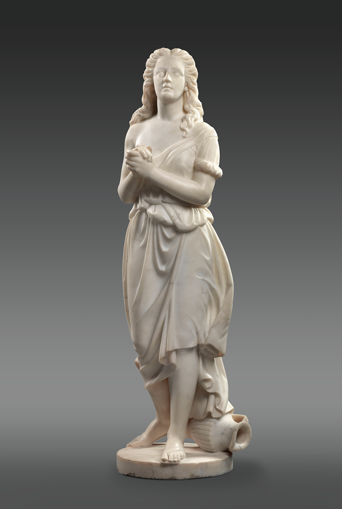 Edmonia Lewis’ marble sculpture of Hagar, the slave of the biblical Abraham and Sarah. She clasps her hands and looks up, her long hair flowing over her shoulders and her loose gown off her right shoulder. At her feet is an overturned water pitcher.