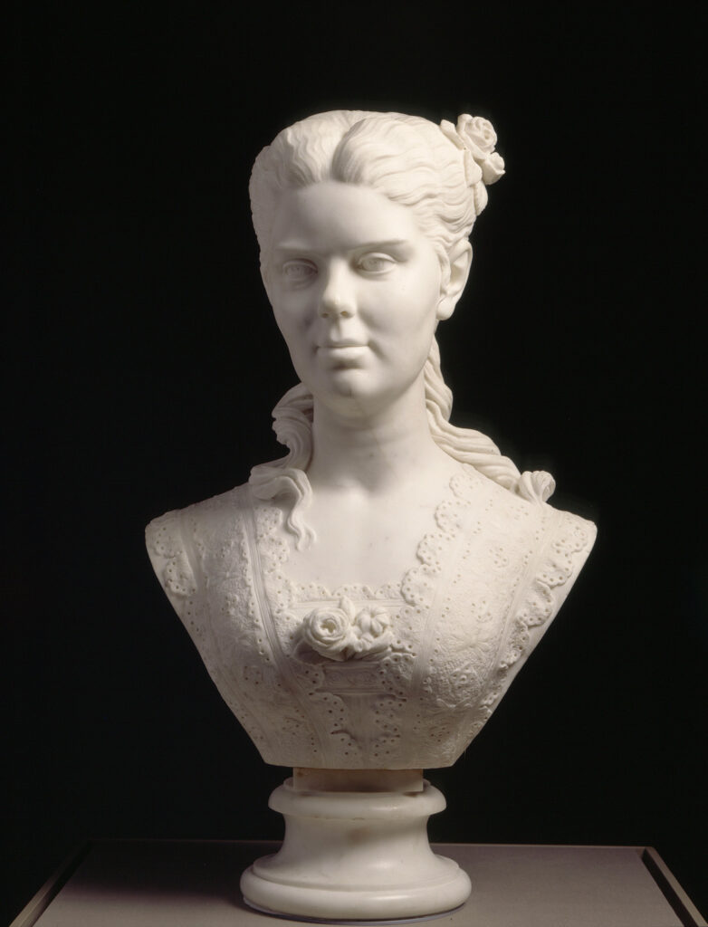 Edmonia Lewis’ “Portrait of a Woman,” a small bust-length sculpture of a woman in a lacy dress ornamented along the bustline. Her hair is parted and pulled back, with curls trailing down her back and shoulders.