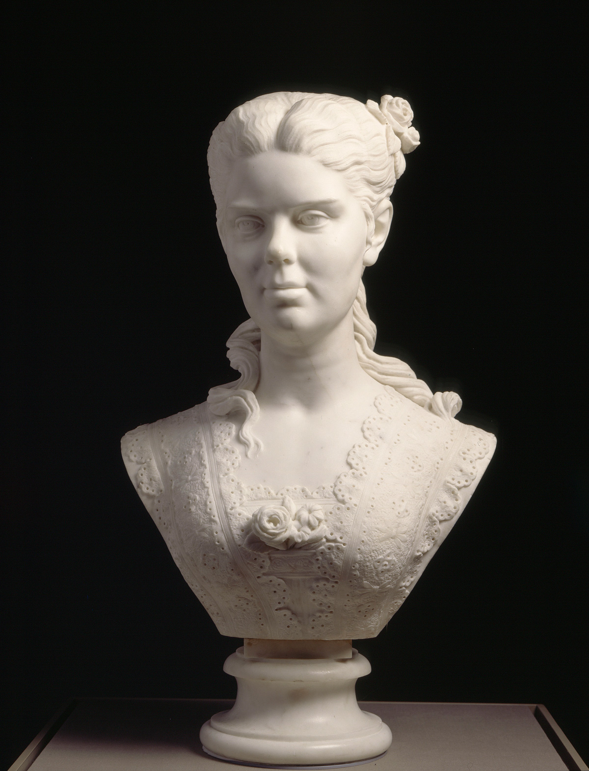 Edmonia Lewis’ “Portrait of a Woman,” a small bust-length sculpture of a woman in a lacy dress ornamented along the bustline. Her hair is parted and pulled back, with curls trailing down her back and shoulders.