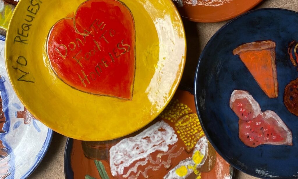 A photograph of several hand-painted ceramic plates. One says "No Request. Donate food to homeless." The others have foods like a baked potato, corn, pumpkin pie and lasagna.