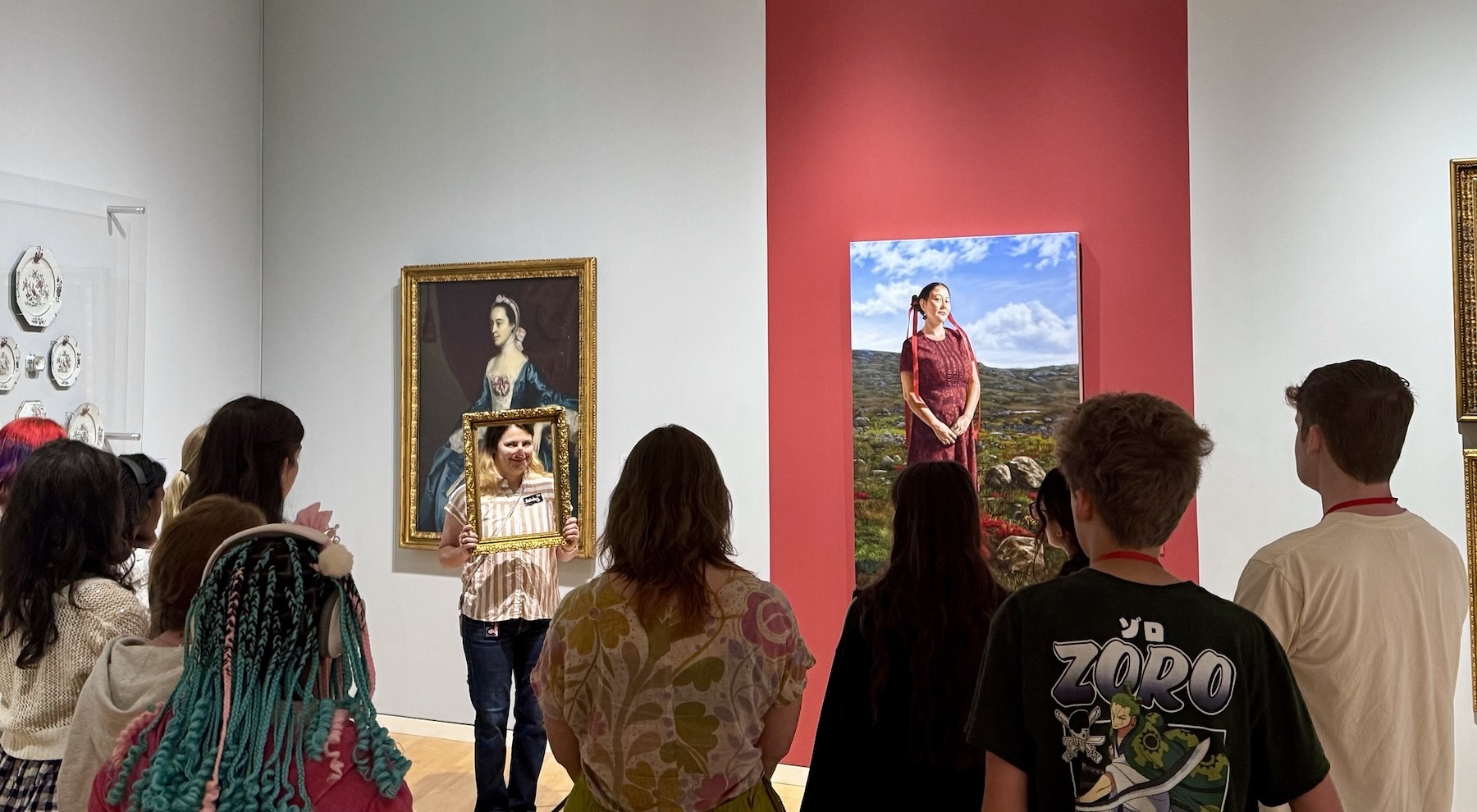 A group of pre-teens listen to a docent, who is holding a frame around her face, in the gallery.